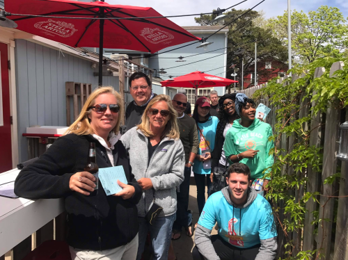 A group of people standing in line outside, smiling and holding drinks, with umbrellas and a sunny atmosphere.