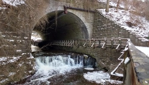 A stone tunnel with a flowing stream and a wooden railing, surrounded by snow-covered banks and trees.