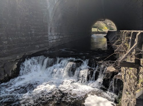 A view of a small waterfall flowing under a stone arch bridge, surrounded by rocks and greenery.