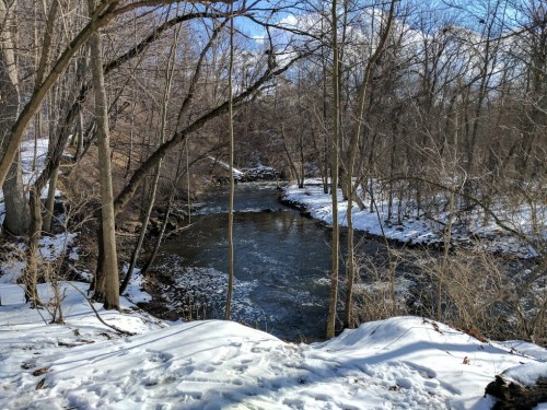 A serene winter scene featuring a river surrounded by bare trees and snow-covered ground under a blue sky.