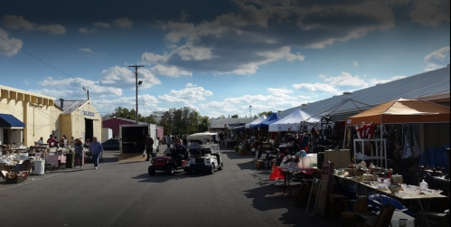 A bustling outdoor market with vendors, tents, and shoppers under a partly cloudy sky.