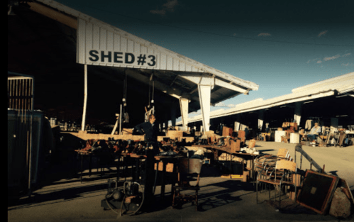 A vendor stands among various antiques and furniture for sale in a bustling market shed.