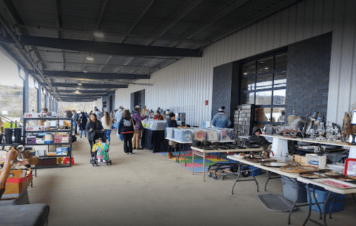 A bustling indoor market with shoppers browsing various stalls and tables filled with vintage items and crafts.
