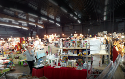 A bustling indoor market filled with various items for sale, with a vendor in the foreground and bright lights overhead.
