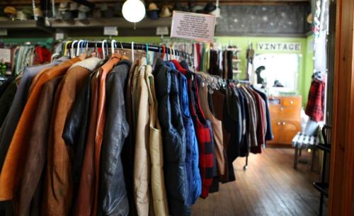 A row of vintage clothing hanging on racks in a cozy thrift store with wooden floors and green walls.