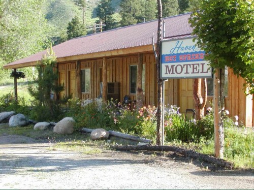 A rustic wooden motel with a sign, surrounded by greenery and flowers, set against a mountainous backdrop.