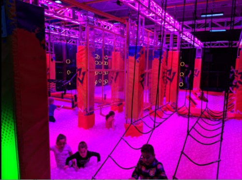 A vibrant indoor play area with colorful structures, ropes, and a foam pit, illuminated by purple lights. Kids are playing.