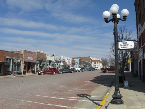 A quiet street lined with shops and parked cars, featuring a vintage lamp post under a blue sky with clouds.