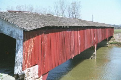 A weathered red covered bridge spans a calm river, surrounded by bare trees and open fields under a clear blue sky.