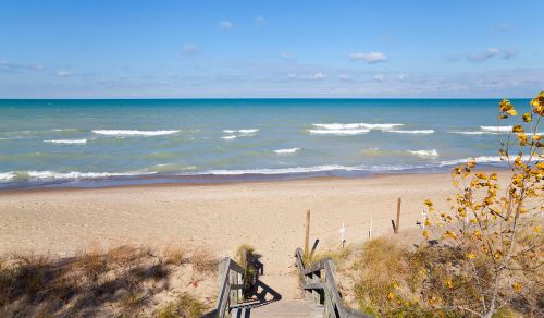 A sandy beach with gentle waves, wooden steps leading down, and a clear blue sky with scattered clouds.
