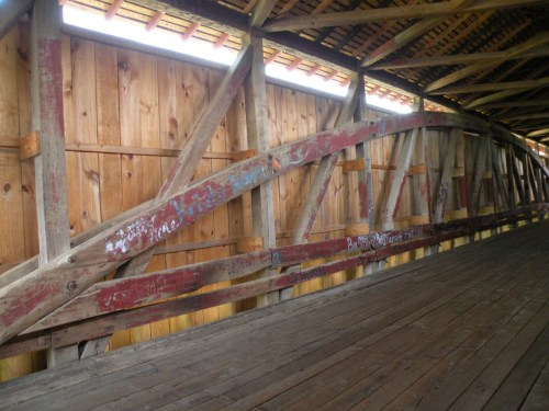 Interior view of a wooden covered bridge, featuring rustic beams and a wooden floor with some graffiti on the walls.