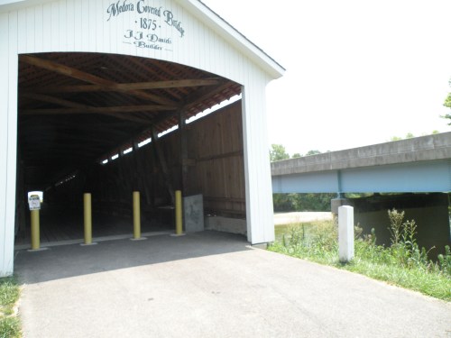 A covered bridge entrance with yellow posts, leading to a river, under a clear sky. Nearby is a concrete bridge.