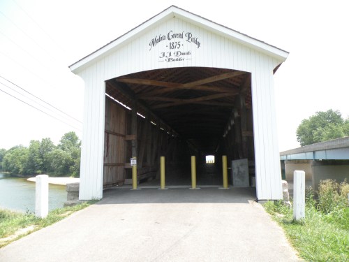 A covered bridge with a white exterior, marked "Alba Creek Bridge 1875," leading to a river and surrounded by greenery.
