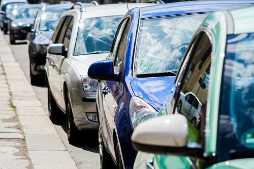 A row of parked cars on a city street, with reflections of clouds visible on the windows.