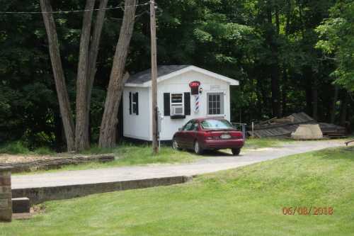 A small white barber shop with a red car parked outside, surrounded by trees and grass.