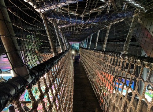 A view down a suspended rope bridge with netting, leading to a colorful area in the background.