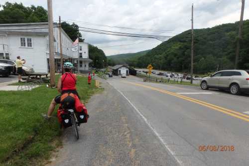 A cyclist in red gear rides along a rural road, with a small town and mountains in the background.