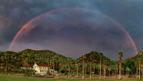 A vibrant rainbow arcs over a mountainous landscape with palm trees and a white house beneath a dramatic sky.