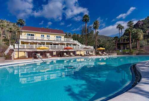 A sunny pool area with lounge chairs, palm trees, and a yellow building in the background against a blue sky.