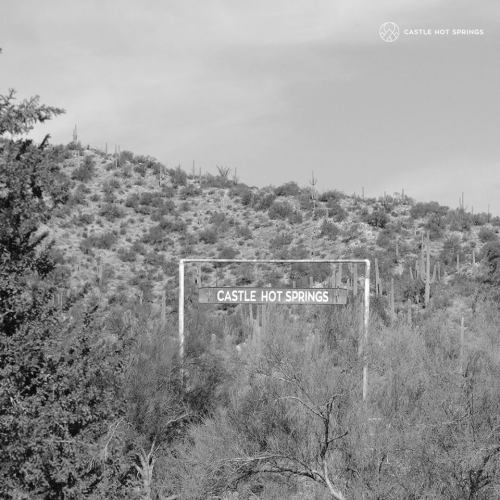 A black and white image of a sign reading "Castle Hot Springs" on a hillside surrounded by desert vegetation.