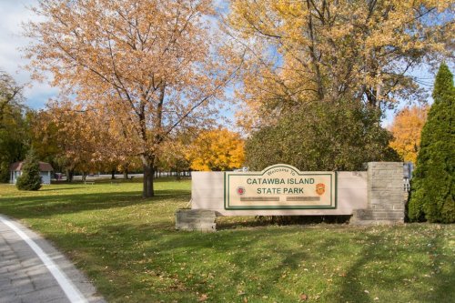 Sign for Catawba Island State Park surrounded by trees with autumn foliage and a paved road.