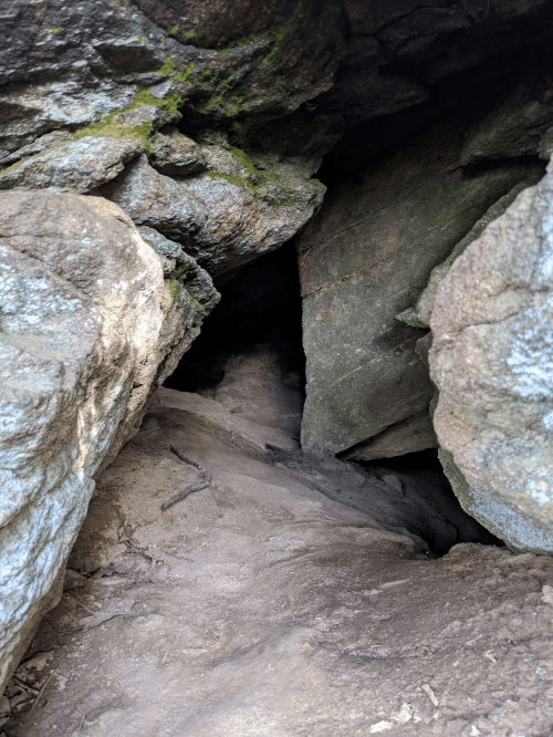 A narrow opening between large rocks leads into a dark cave with sandy ground.