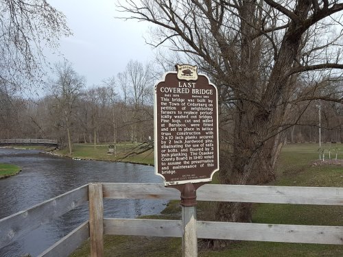 A historical marker about the last covered bridge, with a river and trees in the background.