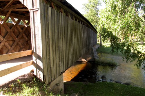 A wooden covered bridge spans a small stream, surrounded by lush greenery and sunlight.