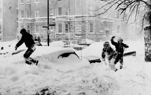 Children playing in deep snow, with snow-covered cars and buildings in the background during a winter storm.