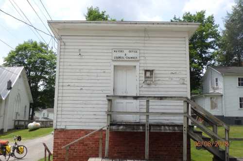 A small, weathered white building labeled "Mayor's Office" with a wooden staircase and surrounding greenery.