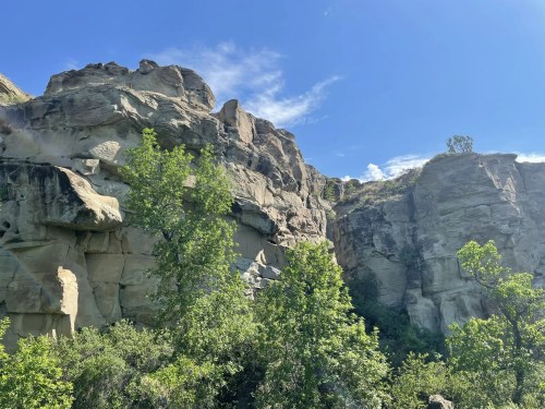 Rocky cliffs rise above lush greenery under a clear blue sky with scattered clouds.