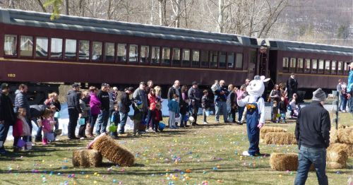 A crowd of families gathers near a train, with a person in a bunny costume and colorful Easter eggs scattered on the ground.
