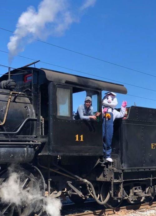 A person and a large bunny mascot wave from the window of a steam train, with smoke billowing from the engine.