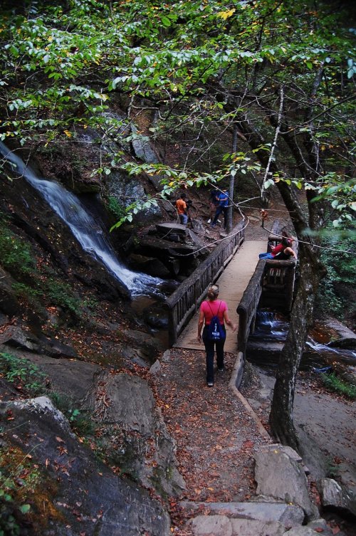 A person walks along a wooden path by a waterfall, surrounded by lush greenery and other visitors enjoying the scenery.
