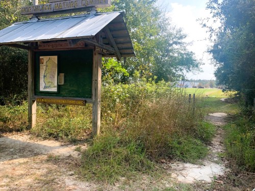 A wooden sign at a nature center entrance, with a path leading into a grassy area surrounded by trees.