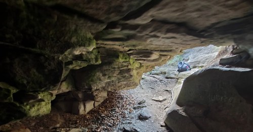 A view from inside a cave, showing rocky walls and a person sitting on the ground outside in a natural setting.