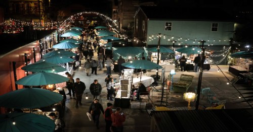 A bustling night market with teal umbrellas, string lights, and people browsing various stalls.