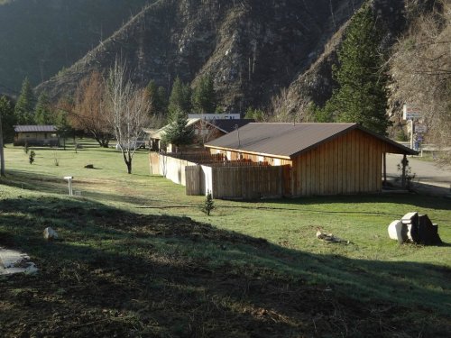 A wooden building surrounded by grass and trees, set against a mountainous backdrop.