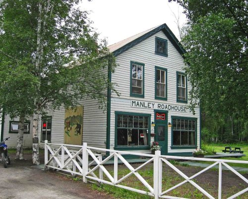 A rustic two-story building with green trim, labeled "Manley Roadhouse," surrounded by trees and a white fence.