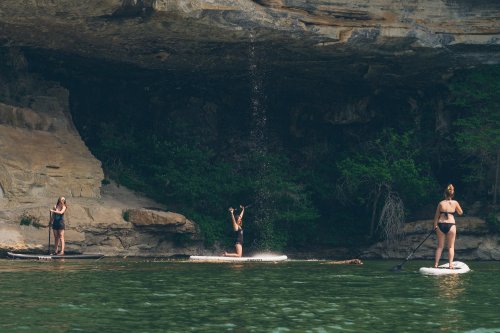 Three women paddleboarding near a rocky cliff, with one splashing water while the others stand on their boards.