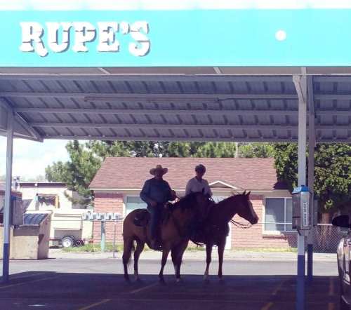 Two people on horseback stand under a sign that reads "Rupe's," with a building and trees in the background.