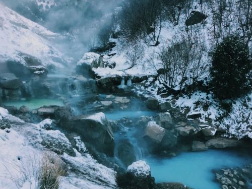A serene winter landscape featuring steaming hot springs surrounded by snow-covered rocks and trees.