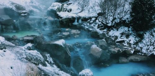 A serene winter landscape featuring a steaming hot spring surrounded by snow-covered rocks and trees.
