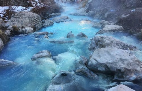 A serene hot spring with steaming blue water flowing over rocks, surrounded by a natural landscape.