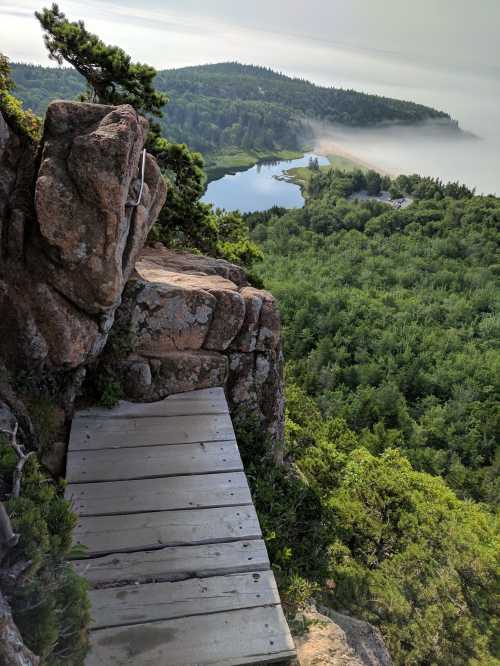 A wooden path leads along a rocky edge, overlooking a lush green landscape and a misty river below.