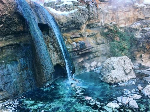 A serene waterfall cascades over rocky terrain into a clear, turquoise pool surrounded by natural stone.