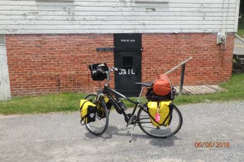 A bicycle with yellow panniers and an orange bag parked in front of a brick wall with a jail door.