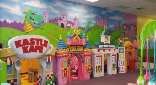 Colorful indoor play area featuring a castle-themed café, salon, and school with vibrant murals and decorations.