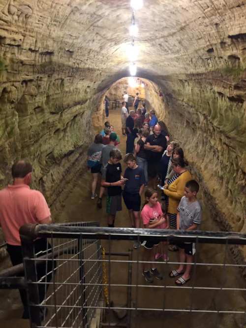 A group of people, including children and adults, explore a lit underground tunnel with rocky walls.