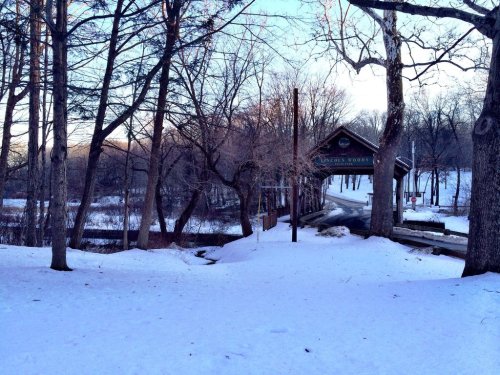 A snowy landscape with bare trees, leading to a wooden bridge over a river at sunset.